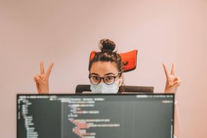 Woman in black rimmed glassed doing victory signs behind a computer display showing code